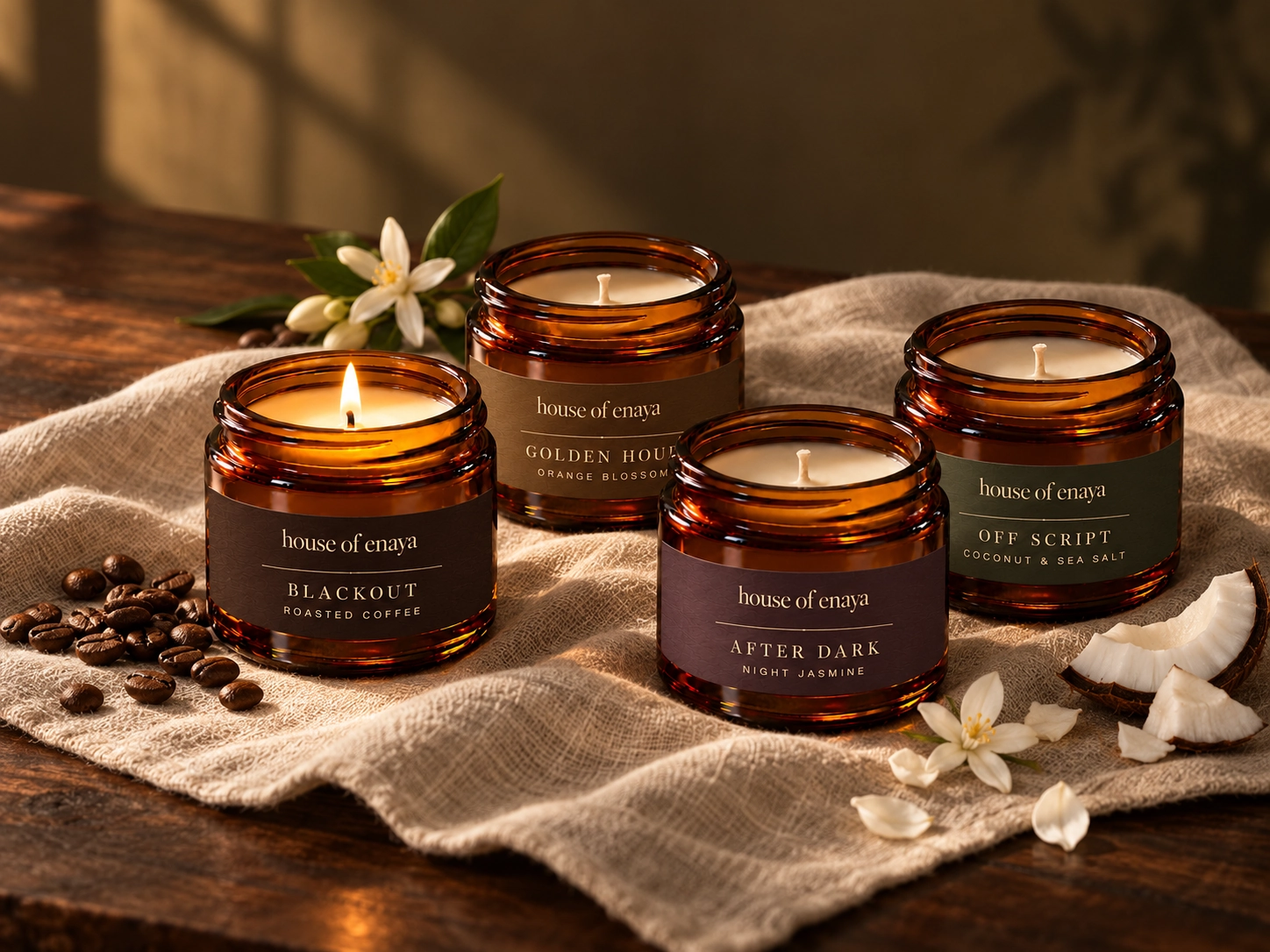 The four House of Enaya 80ml candles on cream linen, with subtle scent botanicals in warm window light